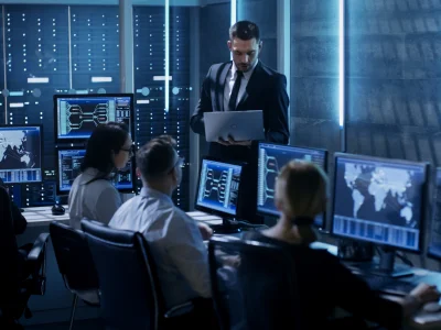 Gruppe vor mehreren Monitoren mit Weltkarten und Codes, Mann im Anzug mit Laptop